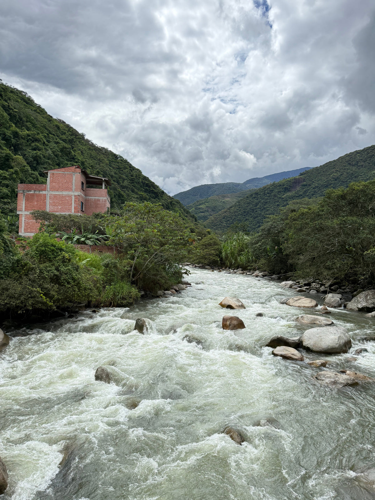 River flowing through the green mountains of Yungas, Bolivia with a small brick house on the left bank of the white fresh water river.