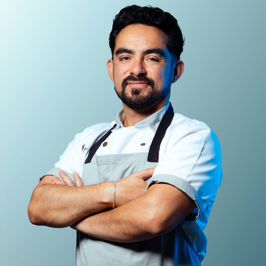 A man, Mario Lima, with a beard and short dark hair, wearing a white chef's jacket and a light gray apron with black straps, standing with his arms crossed against a light blue-gray background.