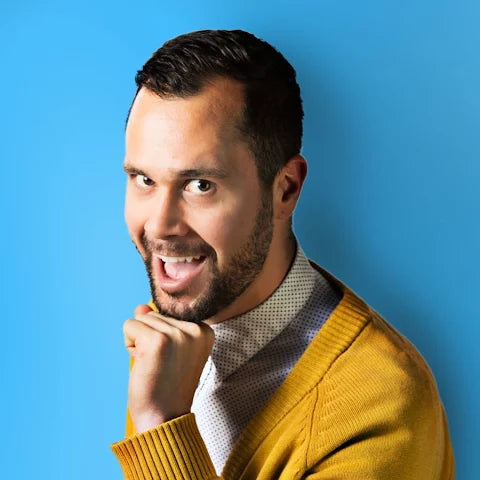 A man, Kyle Privete, with short dark hair and a beard, wearing a mustard yellow knit cardigan over a light patterned shirt, posing against a solid blue background.